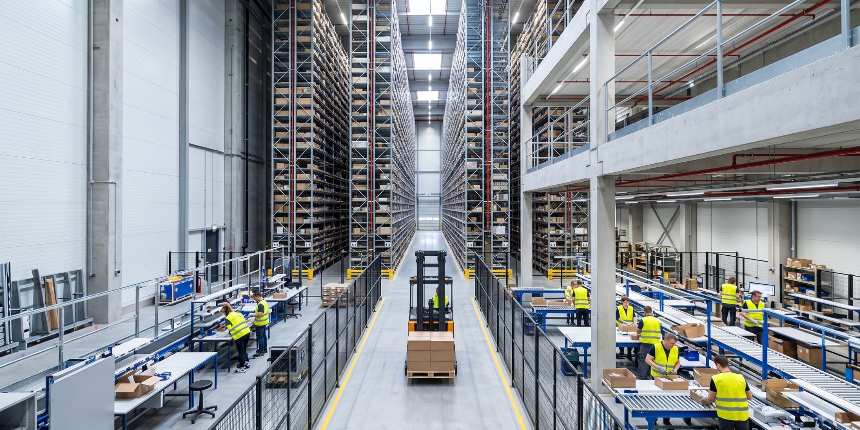 Innenansicht einer modernen Logistikhalle mit Hochregallager, automatisierten Staplern und Mitarbeitern in einem Mezzanin-Bereich. Dynamic der Kontraktlogistik.